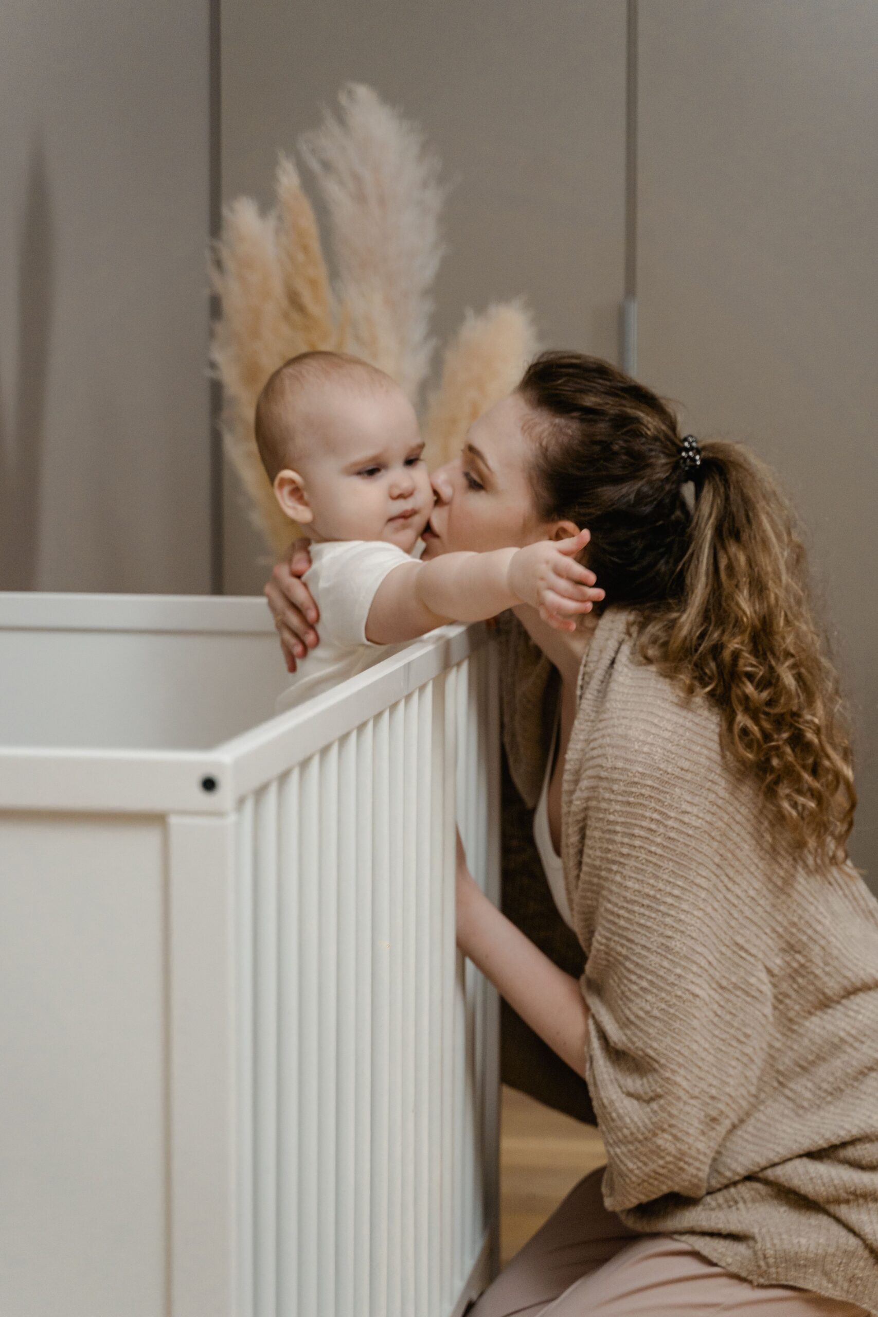 Mother kissing her baby goodnight while leaning over a white crib in a cozy nursery.